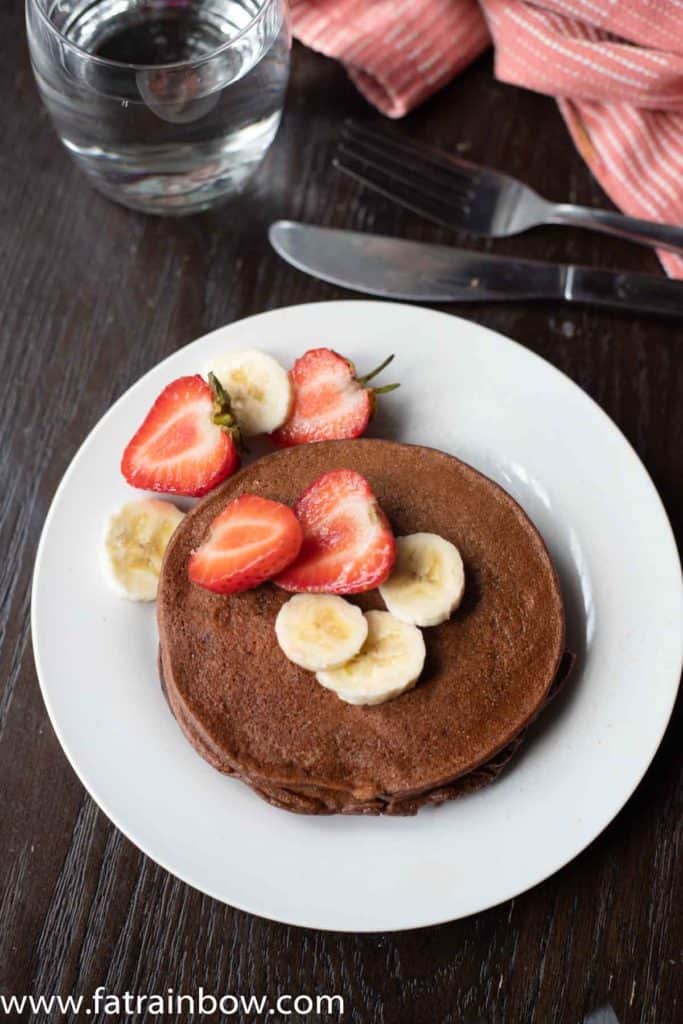 Chocolate protein pancakes served with strawberry slices and banana with napkin and a glass of water in the back