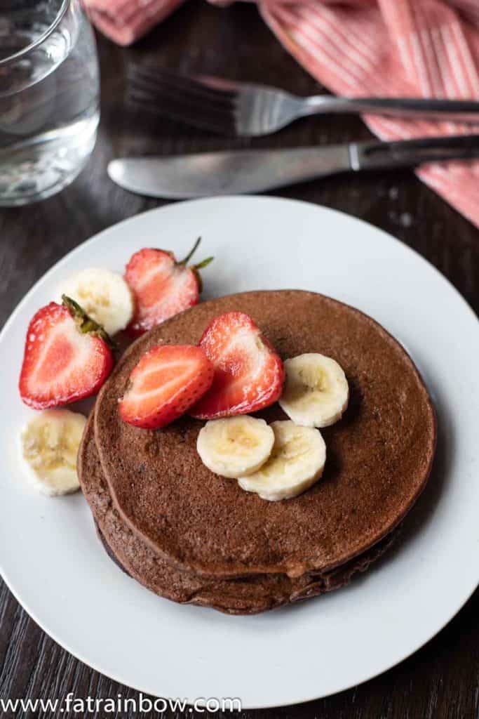 Chocolate protein pancakes served with strawberry slices and banana with napkin and a glass of water in the back