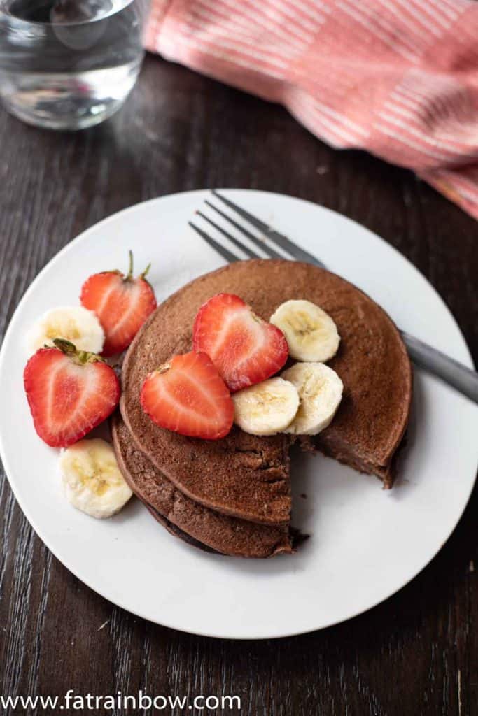 Chocolate protein pancakes served with strawberry slices and banana with napkin and a glass of water in the back