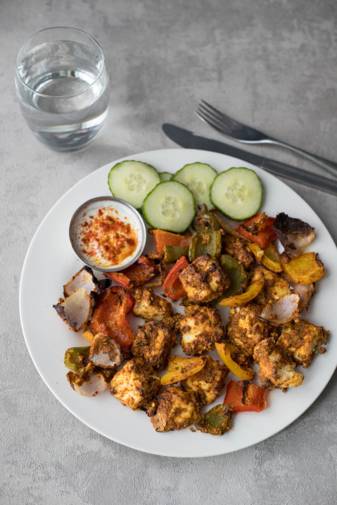 healthy paneer tikka served with dip and cucumbers along with a glass of water and cutlery on side