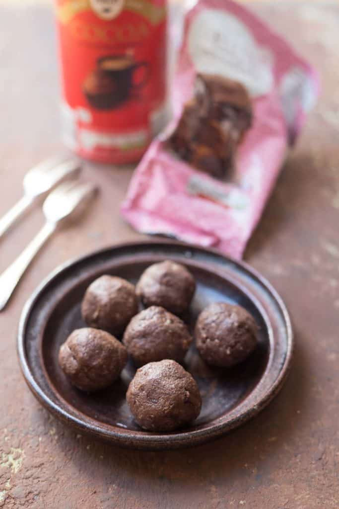 6 Avocado chocolate protein balls served on a brown plate with 2 forks, a pack of chocolate bar and cocoa powder