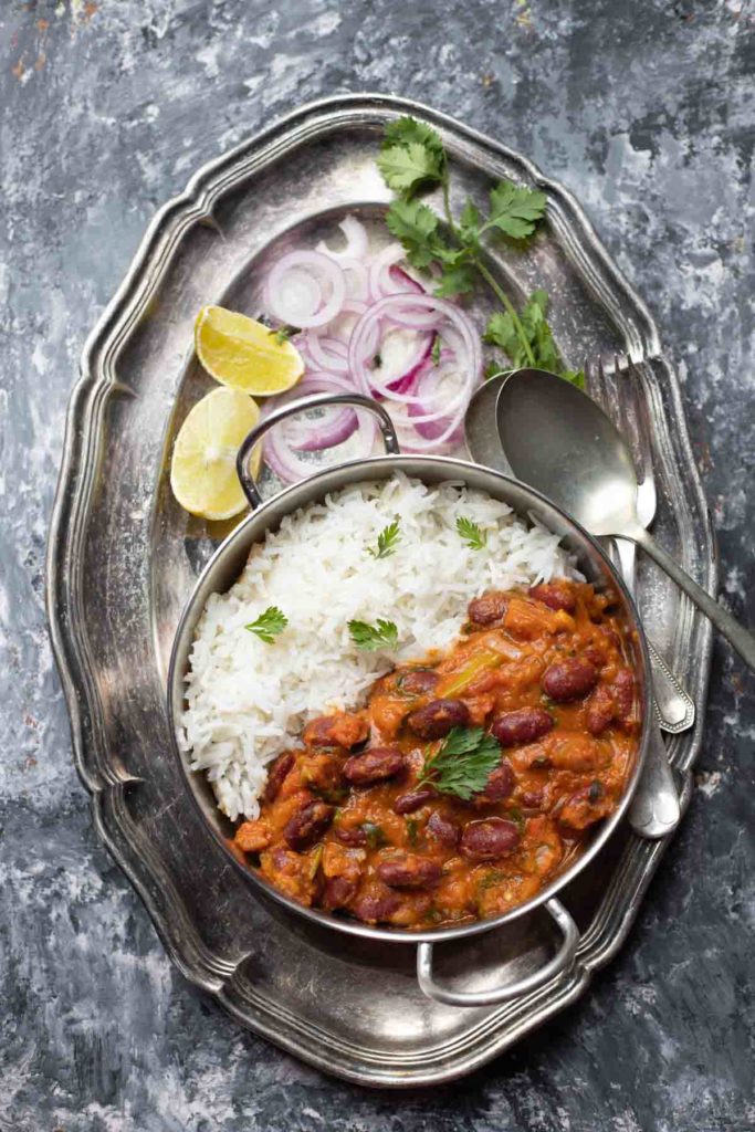  Rajma Masala curry served with basmati rice in a steel kadai placed on a large plate with onion rings, lemon wedges and cutlery