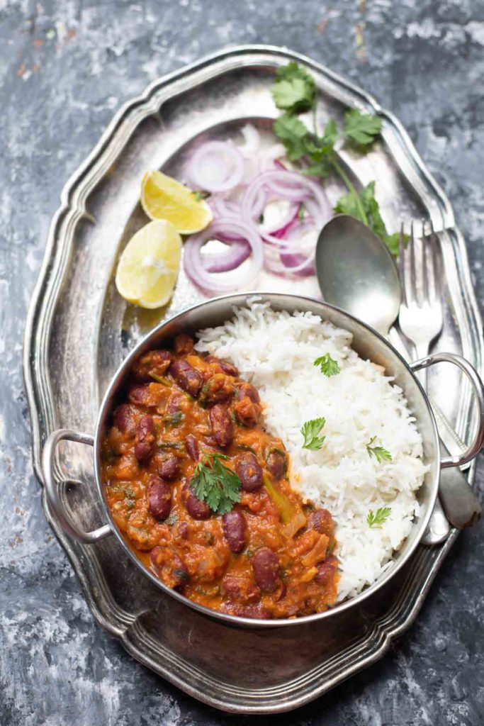  Rajma Masala curry served with basmati rice in a steel kadai placed on a large plate with onion rings, lemon wedges and cutlery