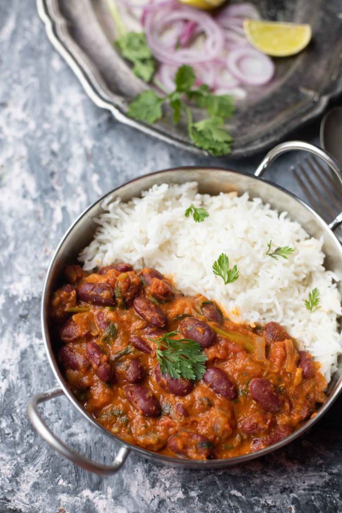  Rajma Masala curry served with basmati rice in a steel kadai placed on a large plate with onion rings, lemon wedges and cutlery