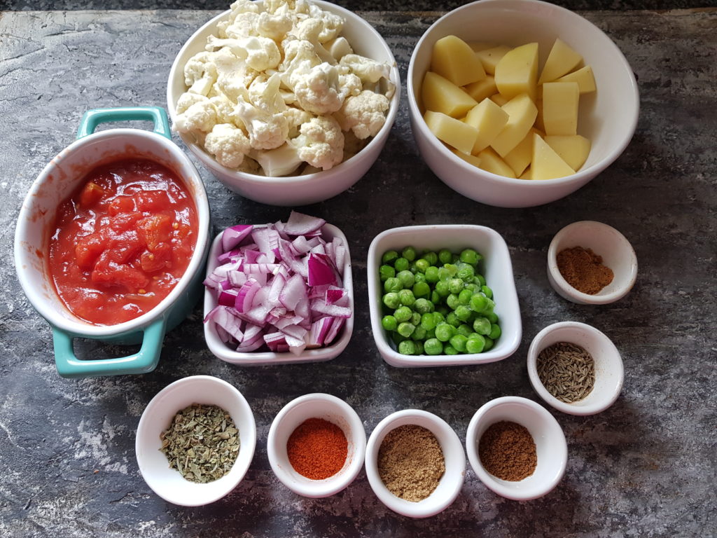 ingredients for making aloo gobi, tomatoesm potatoes, cauliflower,onions,peas, spice powders,cumin seeds