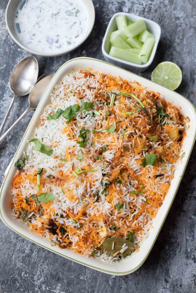 hyderabadi dum biryani in a oven proof dish , cucumber salad, raitha, lime wedge, spoons