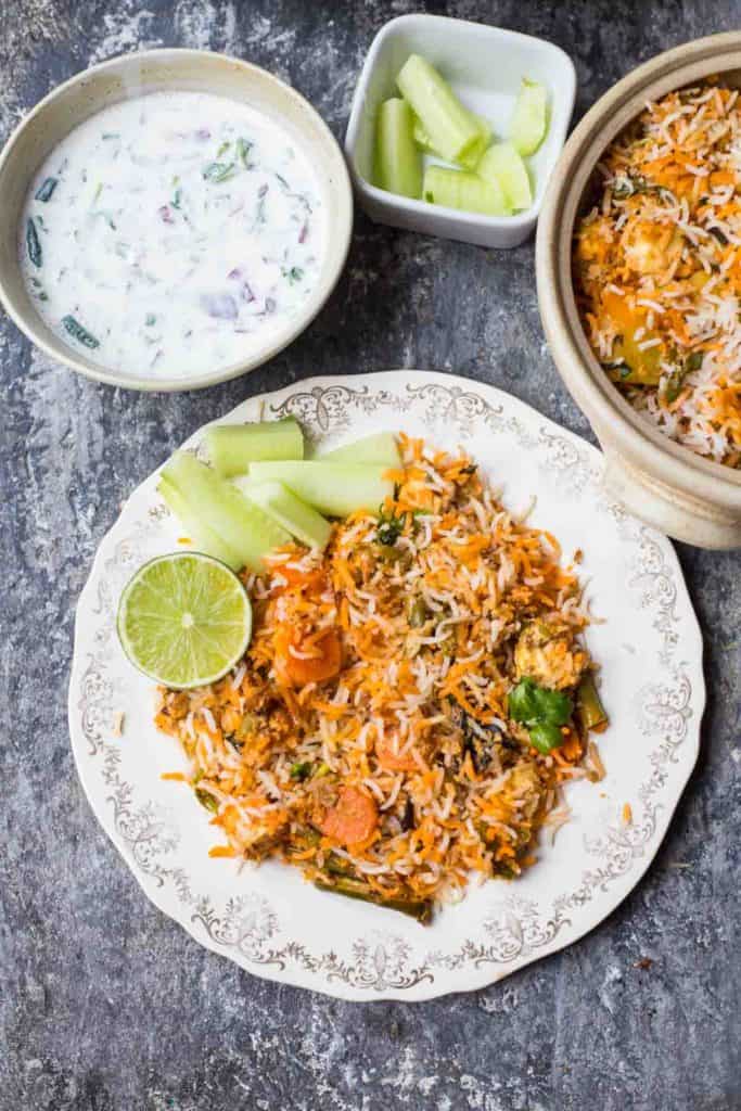 hyderabadi dum biryani in a plate , cucumber salad, raitha, lime wedge, spoons