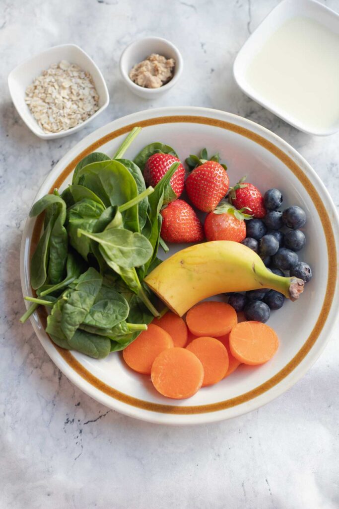 a plate loaded with berries, banana, carrots,spinach and a bowl of oats , milk and peanut butter