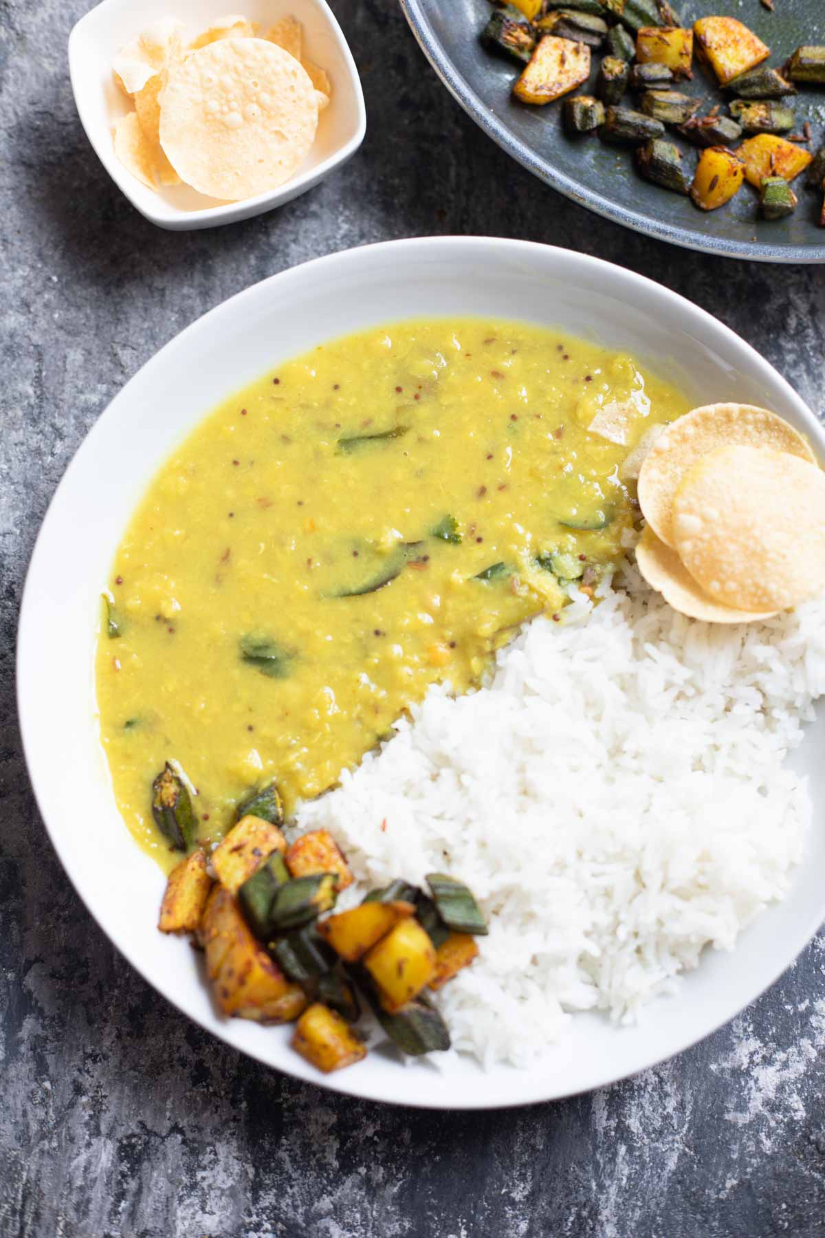 raw mango dal served with rice and aloo bindi stir fry in a white plate, papads in background