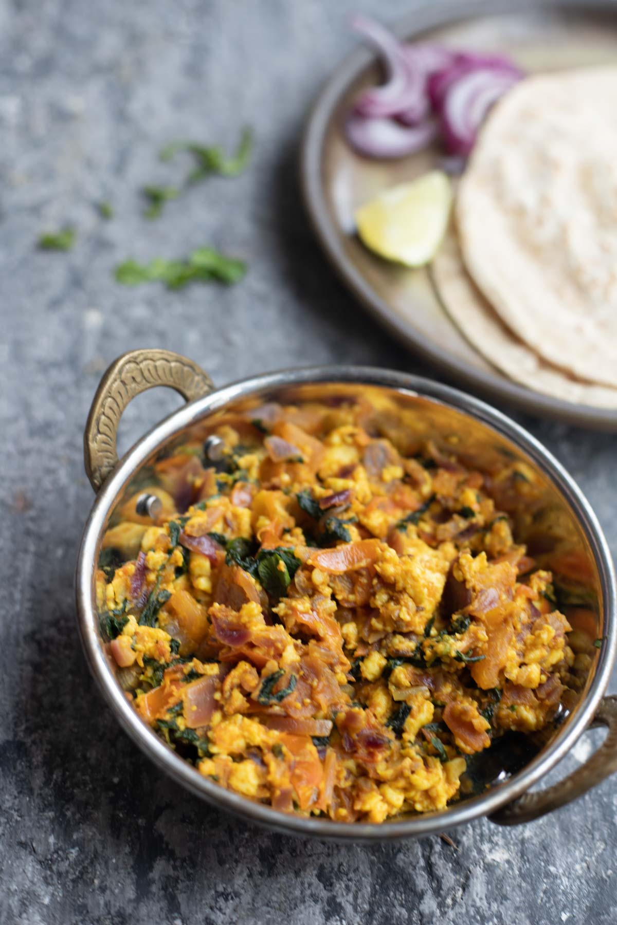 methi paneer bhurji in a steel kadai with a plate of rotis , onion and lemon salad in the background