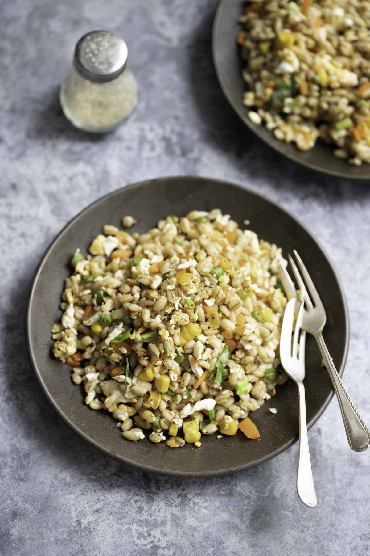 two plates of barley fried rice with a ground pepper jar in the background