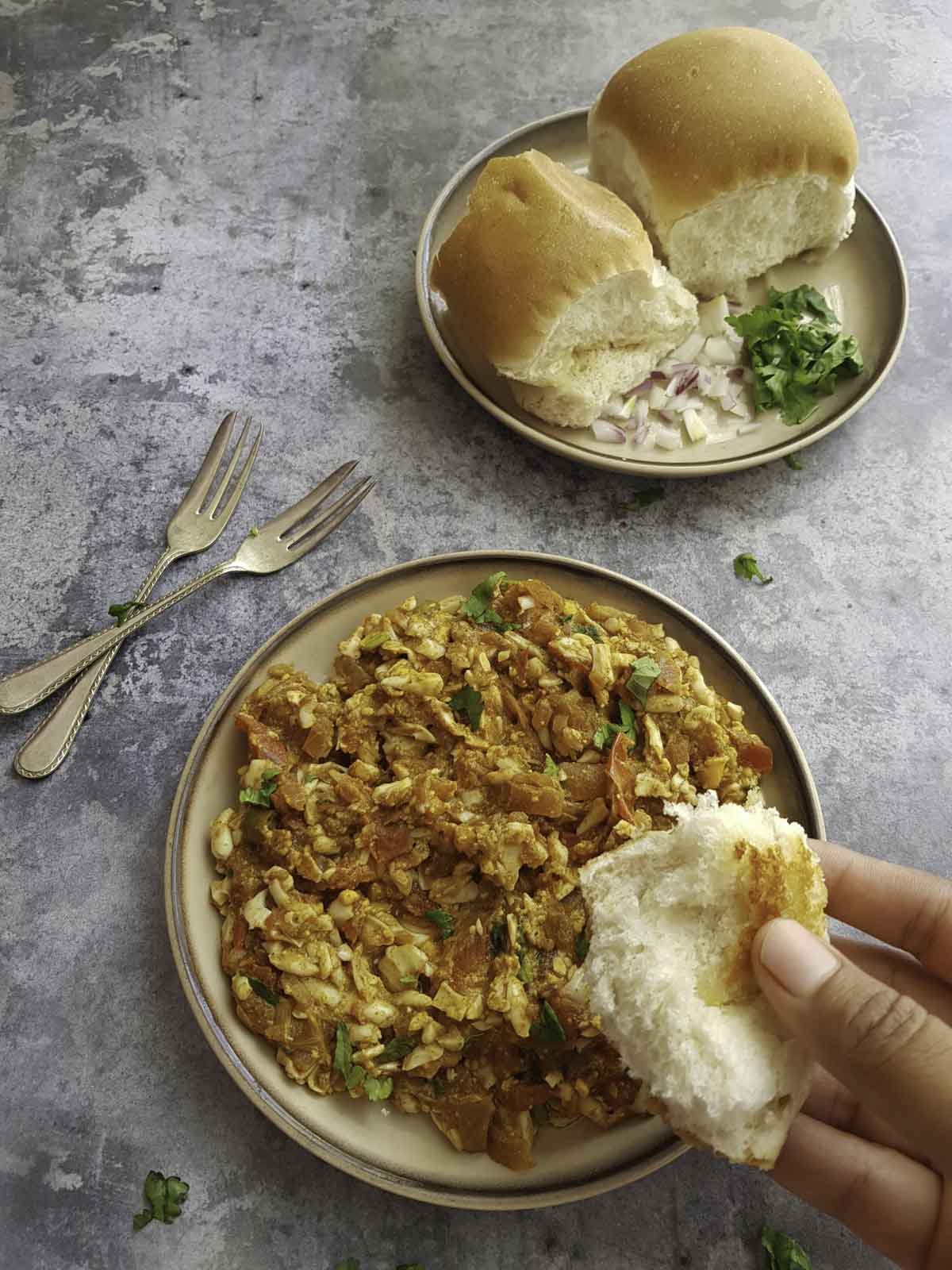 a hand  dipping bread piece into keema egg , two forks and a plate of pav breads on the side.