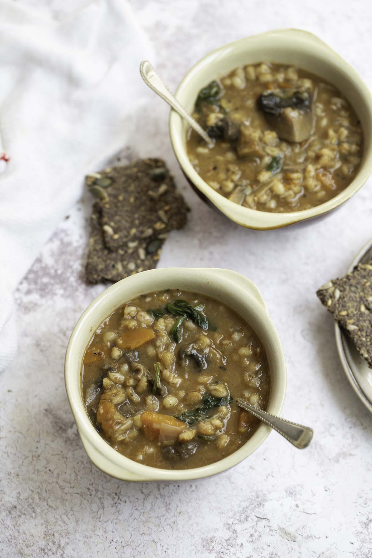 two bowls of vegetable barley soup with spoons, crisp bread on the side