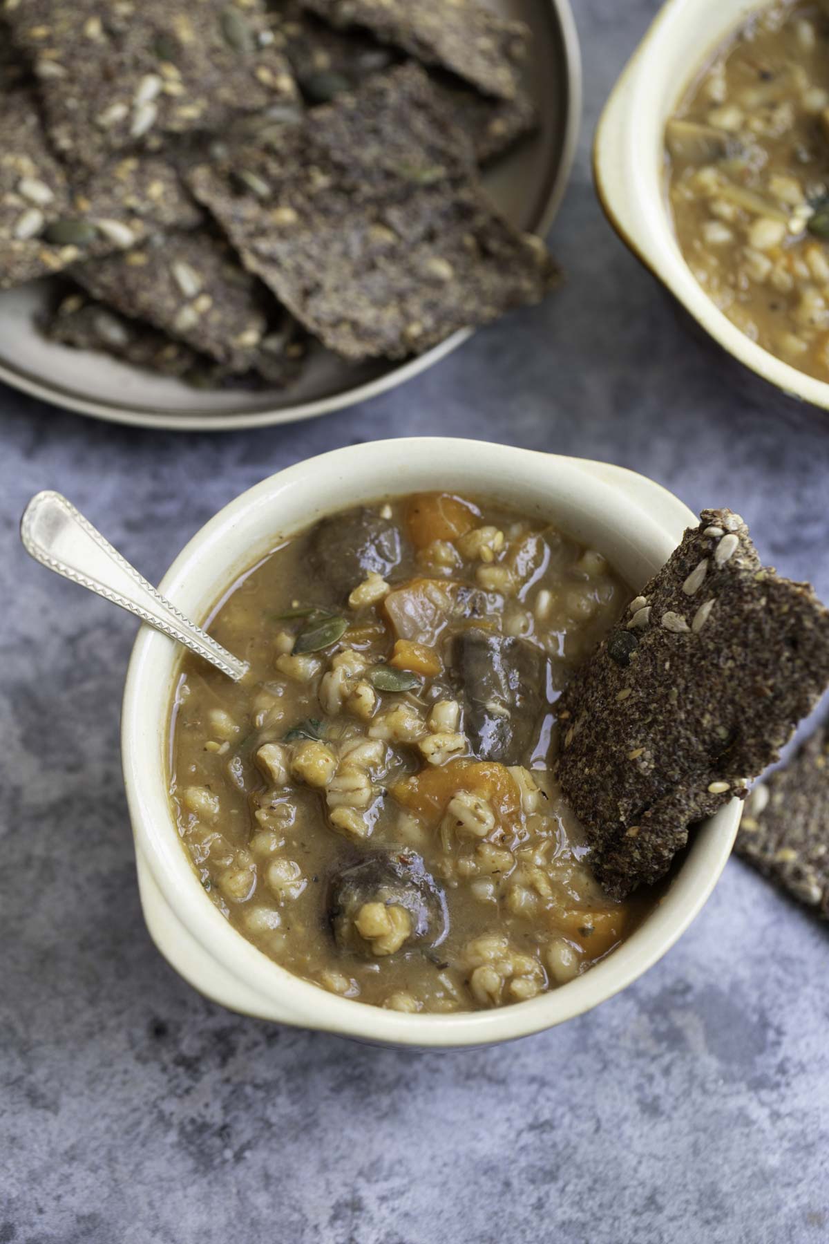 a bowl of vegetable barley soup with a crisp bread and silver spoon